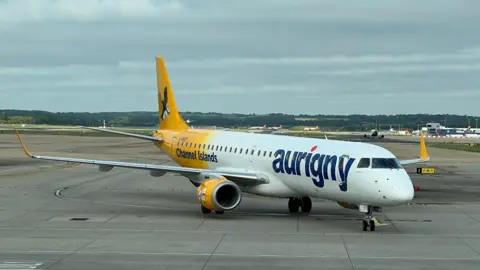 BBC A shot of an Aurigny aeroplane at an airport. It is mainly white with the company branding on it with the back of it yellow. It has Aurigny and the Channel Islands labelled on it. 