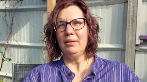 A woman with wavy brown bobbed hair looks just to the side of the camera with a slight frown. she is wearing dark-rimmed glasses and a blue and white striped blouse.