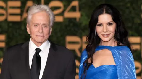 Getty Images Michael Douglas and Catherine Zeta-Jones are standing next to each other facing towards the camera smiling. Catherine is wearing a blue dress and Michael is wearing a black suite with a white shirt and black tie. 
