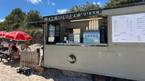 A picture of the beach cafe in Grouville in a port-a-cabin style building. Some picnic benches with umbrellas are pictured alongside on the sand.