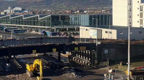 An aerial view of the  The Boyne Bridge in Belfast in January 2025.  The bridge is closed off with barriers during demolition work.  A yellow digger is in the foreground and Grand Central Station is in the background. 