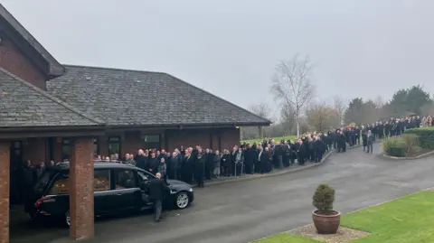 A long queue of people, all in black, on the pavement as a hearse with a coffin backs up into the crematorium entrance