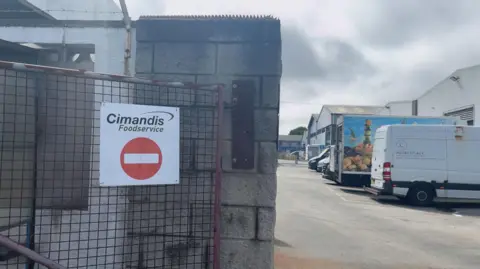 BBC Photo of a Cimandis Foodservice 'no entry' sign on a gate in the foreground to the warehouse. In the background there are vehicles parked. 