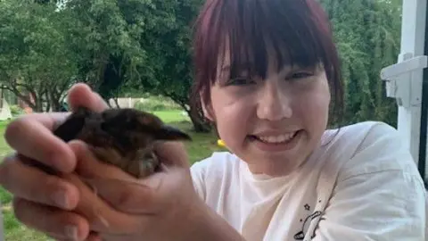 Family handout A young girl holding a small chick and smiling at the camera. She is wearing a white top and has fringe over her face
