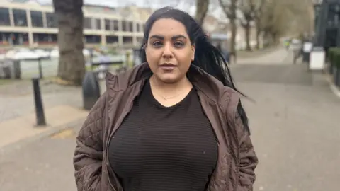 Meena Najmi standing in front of Bristol Harbour, opposite V-SHED. She has dark hair and is wearing a brown top with a brown coat.