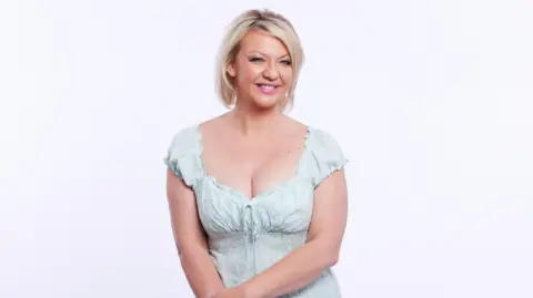 Becky is smiling- she has short blonde hair and is wearing a dress.
