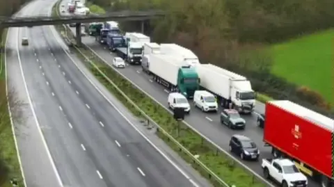 A six-lane motorway. The side to the right of the screen is full of stationary cars and lorries. The side to the left is empty of traffic. Green fields are visible to the right, and a pedestrian bridge can be seen in the distance. 