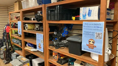 A wooden shelving unit full of various DIY equipment, toolboxes, vacuums, carpet cleaners, cooking equipment, and camping gear. There are three blue posted taped to the shelves displaying an appeal for donations to fund the scheme. 