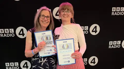 Two women are standing side by side holding up certificates and a glass award. They are both smiling. They are in front of a black back drop with the BBC Radio 4 logo printed on it.