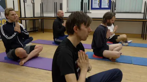 John Fairhall/ BBC Multiple men are sat on yoga mats, the majority have their eyes closed and their hands together in a praying motion. 