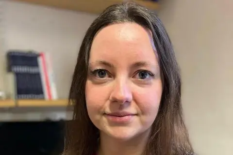 Dr Nikki Rutter has long brown hair with a centre parting and blue eyes. She is looking directly into the camera in front of a bookshelf in an office.