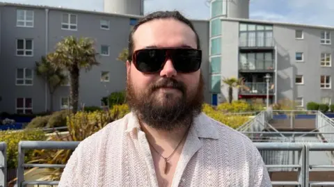 Curtis pictured with apartments visible in the background, alongside some foliage. He is wearing dark glasses, and has a brown beard