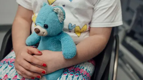 Getty Images A small child is sitting on a chair. They are wearing a white top with yellow and blue hearts on it. They are wearing white, pink and blue leggings and are holding a blue bear on her lap