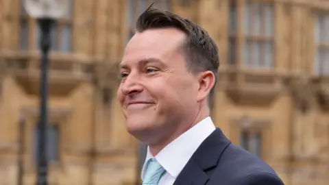 PA Media James McMurdock is wearing a blue tie, white shirt and navy blue suit jacket. He is side-on to the camera, smiling, and the Palace of Westminster appears to be out of focus in the background.