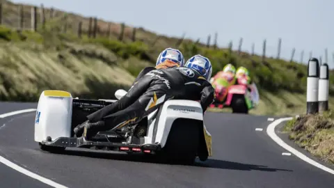 IOM TT A sidecar outfit from behind on a section of the Mountain road. Another outfit is ahead on the road rounding a bend.