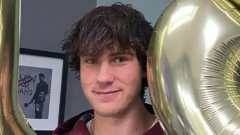 A man with dark hair is looking at the camera. A grey background is behind him on the left.