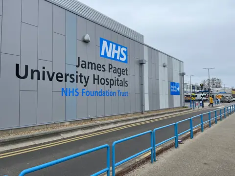 Andrew Turner/BBC Exterior image of the James Paget Hospital in Gorleston. The large grey building has a blue guardrail around it and the name of the building is written on it in large letters