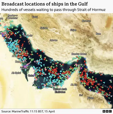 A satellite image of the Strait of Hormuz with ships marked with arrows. It shows hundreds of ships clustered by the strait waiting to pass. It is timestamped to 11:15 BST on 15 April