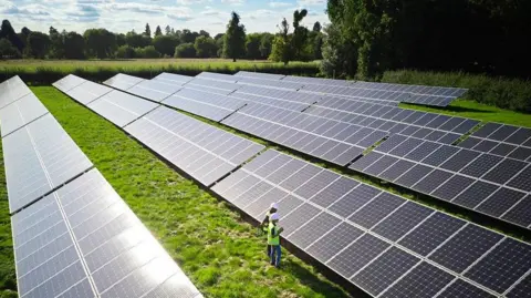 Five rows of large solar panels in a field. Two people wearing high visibility jackets and white hard hats are stood in front of the panels. Trees and hedgerows surround the field.