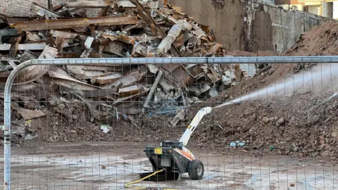 Ollie Conopo/BBC A small black and white robotic machine spraying water on rubble, with a high pile of rubble in the background.