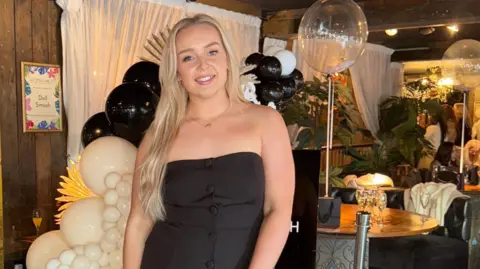 Jess Thorpe Jess Thorpe posing for the camera in a black strapless dress. She has long blond hair and is standing in front of a black and white balloon display in a place that looks like a restaurant.