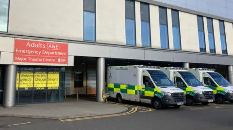 Three ambulances parked outside the Queen Elizabeth University Hospital. A sign of the building reads Adults A&E, emergency department and major trauma centre.