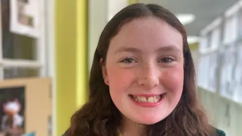 BBC Sophie has long brown wavy hair and a middle parting. She has brown eyes and a gap between her two front teeth.