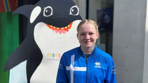 A young female athlete in a brightly coloured blue tracksuit smiles alongside a cutout of Ola the Orca