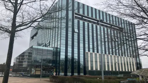 Nadia Lincoln/LDRS A large glass building with a few windows open, in the foreground are a couple of trees 
