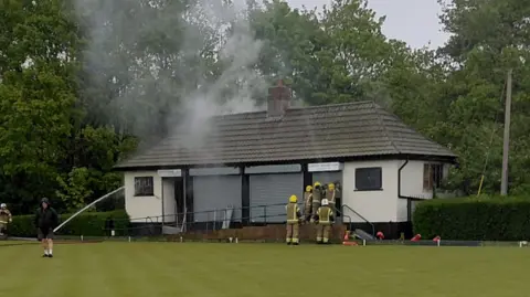 Paul Donaghy Firefighters stand on the bowling green and also on a concrete ramp leading into the club house as the building is ablaze. Smoke is rising from the roof, which has been partially damaged.