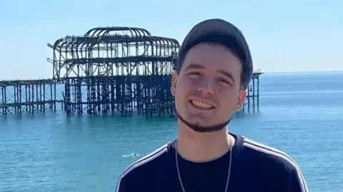 A man wearing a dark top and baseball cap stands in front of the remains of Brighton West Pier, with the sea in the background.