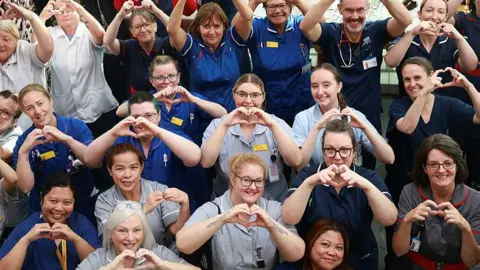 The Royal Wolverhampton NHS Trust People dressed in NHS nurse outfits making heart gestures with their fingers at the camera. Most of them are female and dressed in uniforms ranging from grey to deep blue