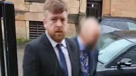 Kevin Montgomery - a man with ginger hair and a beard, arriving at court. He is dressed in a suit with a blue tie. Another man, whose face has been blurred, is walking beside him. 