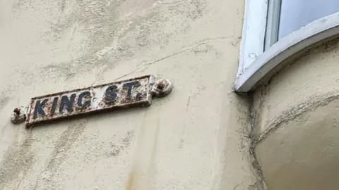 A rusty road sign for King Street bolted to a dirty beige wall.