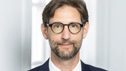 Lampard Inquiry Nicholas Griffin KC, wearing a dark blue suit and white shirt. He has round dark-rimmed spectacles and short beard. He has short dark hair and is stood in front of a light blurred background. He is looking directly at the camera.