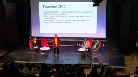 A panel discussion taking place in an auditorium. Four speakers are seated on stage in red chairs, with one person standing and speaking to the audience. Behind them is a large projection screen displaying a slide titled “Timeline CICC,” which lists key dates and events related to the Intergenerational Climate Crimes Act. The audience is seated in rows facing the stage.