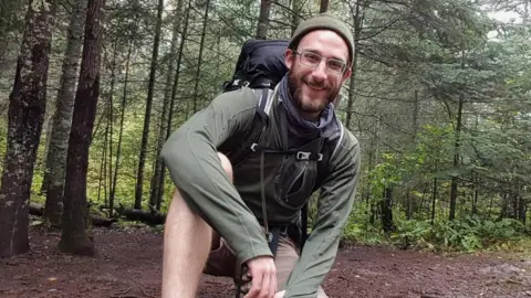 Alex Pretti smiles and kneels on one knee in a wooded area. He wears a green hat, green long-sleeved top, brown shorts, and a black hiking backpack. He has brown facial hair. It is daytime.