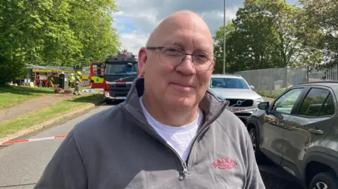 Alex Pope/BBC A man wearing a zipped jumper is stood on a closed road with fire engines parked behind him.