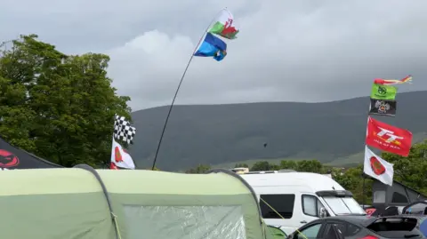 Flag poles with a range of colourful flags blow in the wind. You can see a green tent in the foreground and vans and cars parked up nearby.