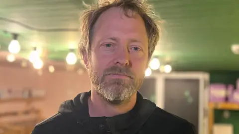Kaspar Wimberley standing in the SCOOP Sustainable Co-operative store in St Lawrence. He is wearing a black hoodie and has gray hair and a beard.