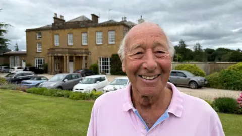An older man in a pink T-shirt smiling at the camera. There's a grand stately home in the background with a car park in front.