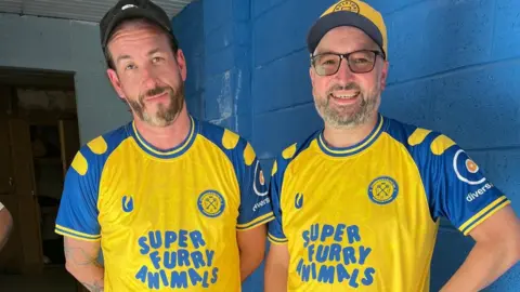 Connor Cupples and Matt Jarrett stood in the new team kit for the men's team, which is yellow with blue short sleeves and super furry animals written on the front also in blue. 