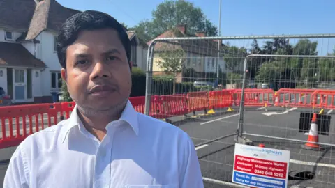 Tom Jackson/BBC Delowar Hussain has short brown hair and is wearing a white shirt. He is standing in front of a sinkhole which has a metal fence around it. 