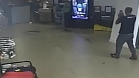 CCTV image of a man wearing a black t shirt and trousers walking towards a machine with a lit up screen inside a building. Racks and shelves can be seen on the left.