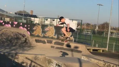 Jake Adams Jake, who wears black trainers, a black cap and black shorts, and a white t-shirt, does a trick mid-air while skating in the graffiti-covered skate park. 