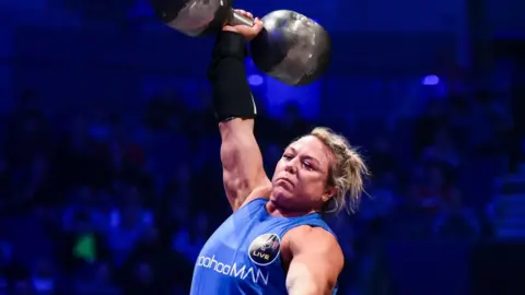 Giants Live A woman in a blue vest lives a large dumbbell above her head with one arm