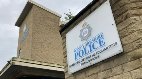 White sign on a pale stone wall with the words West Yorkshire Police Bradford headquarters written on it in black