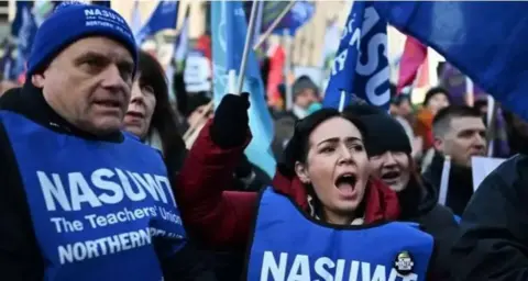 Charles McQuillan A man and woman are taking part in industrial action. They are wearing blue bibs with 'NASUWT' printed on the front. There are surrounded by other people, some are waving coloured flags. 