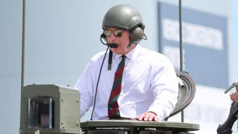 King Charles has the top half of his body sticking out of the top of a tank, he is wearing a white shirt with a red and green tie and some sunglasses. He is also wearing a green helmet with ear defenders and a microphone.