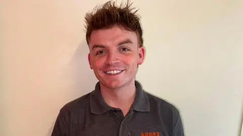 Caoimhinn Campbell has long brown spikey hair and is wearing a grey shirt.